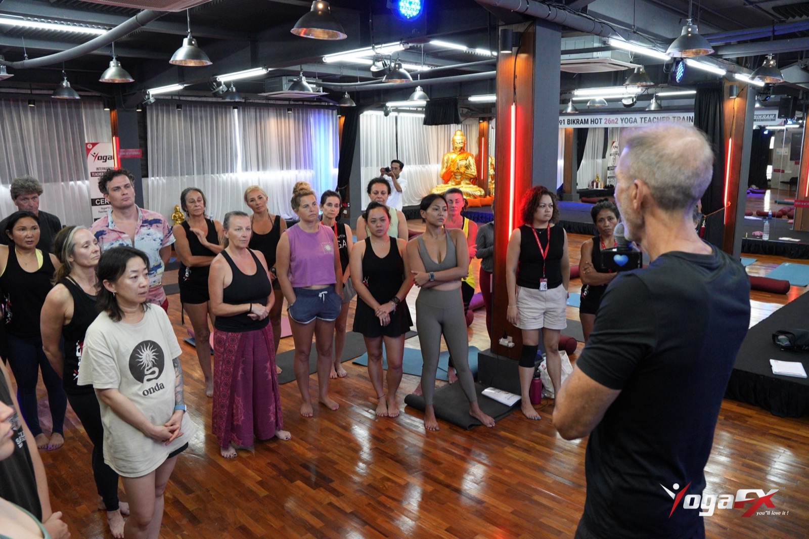 A teacher training cohort standing together in the Bali studio while Mr Ian Terry briefs them before practice