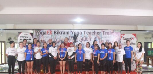 A YogaFX teacher training intake photographed together on stage beneath the Bikram Hot 26 and 2 banner in Bali